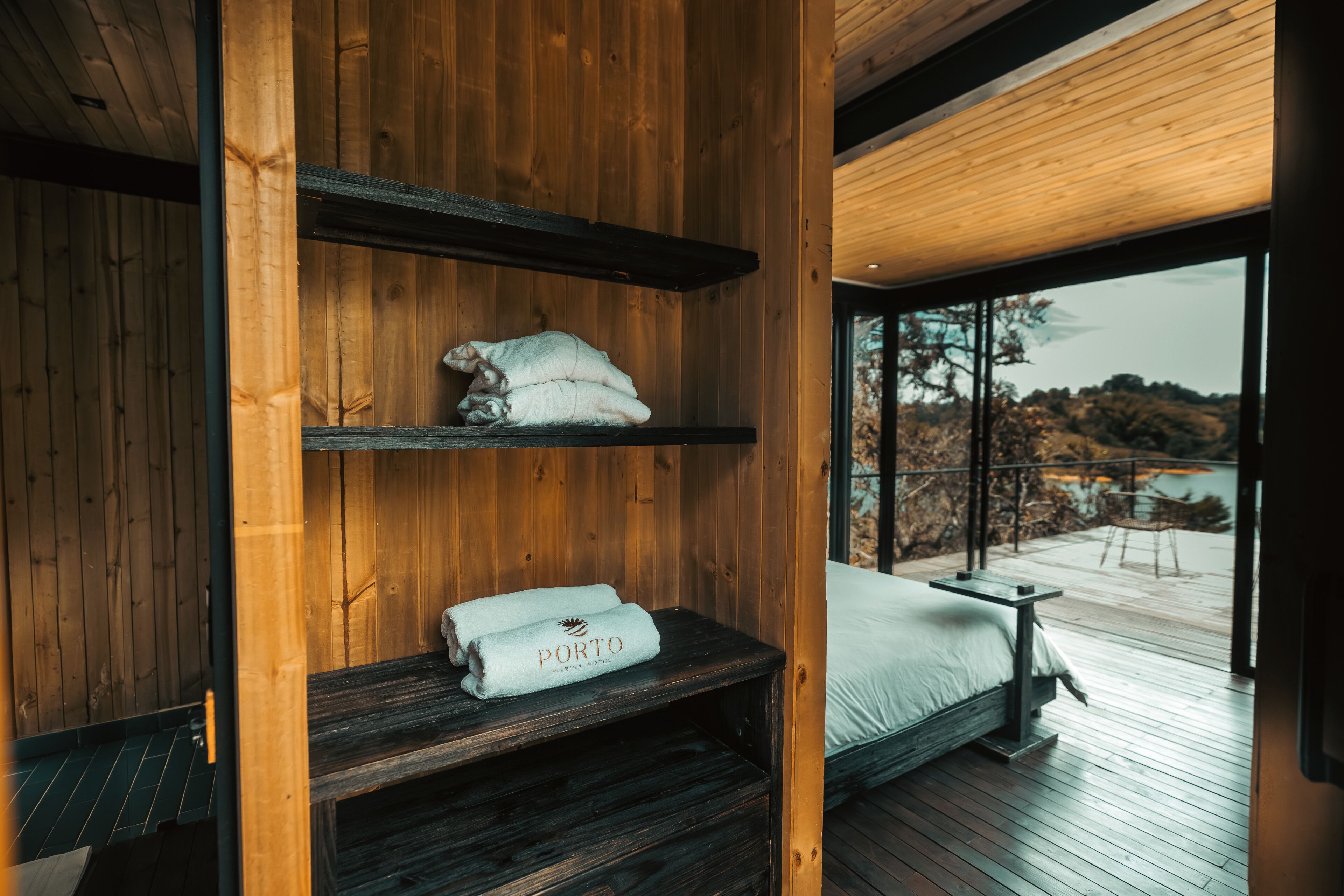 A wooden cabin interior with a bed, a shelf with towels, and a view of the outdoors.