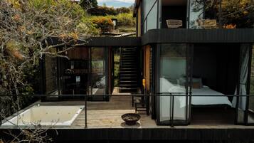 Una casa moderna con un dormitorio con paredes de cristal, una terraza de madera y un jacuzzi.