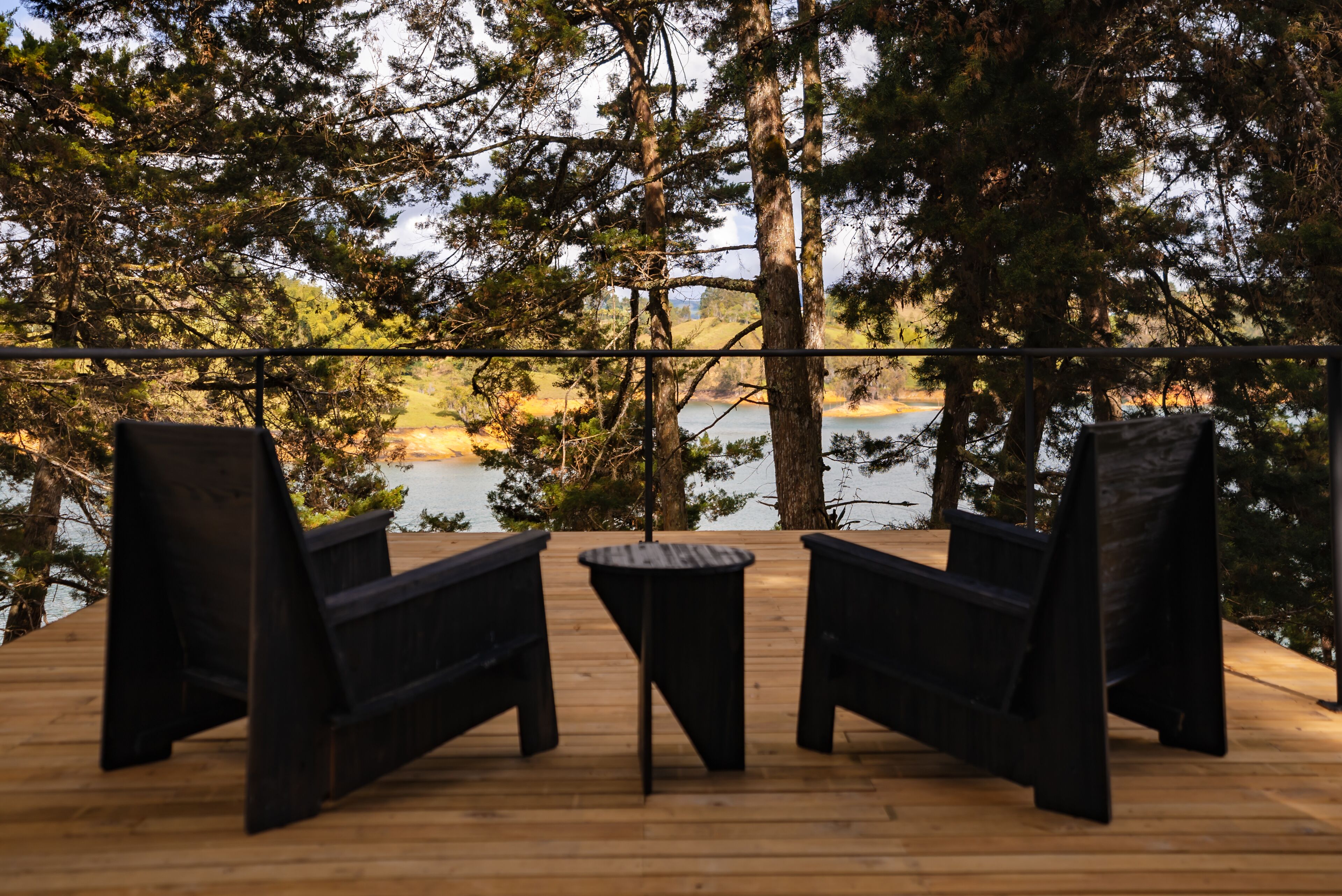 A wooden deck with two black chairs and a small table, overlooking a body of water and surrounded by trees.