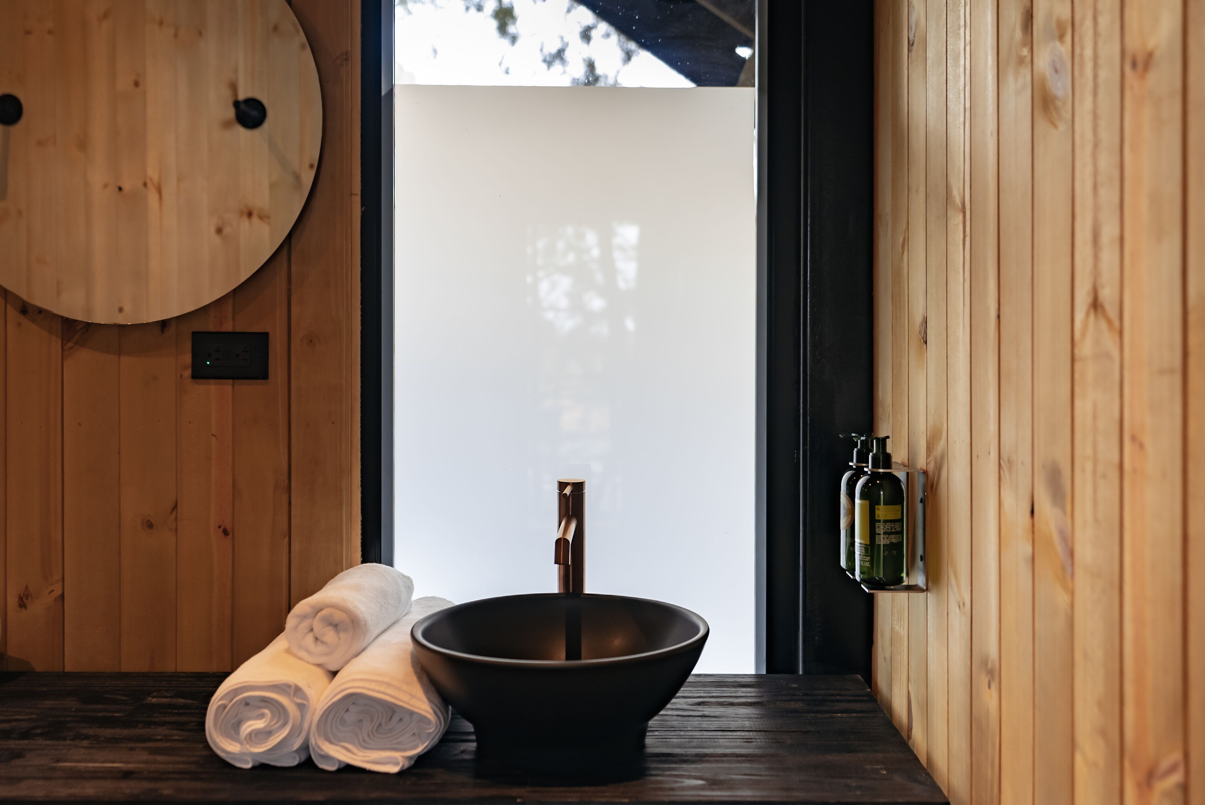 A wooden room with a large round mirror, a black basin, and rolled towels on a wooden bench.