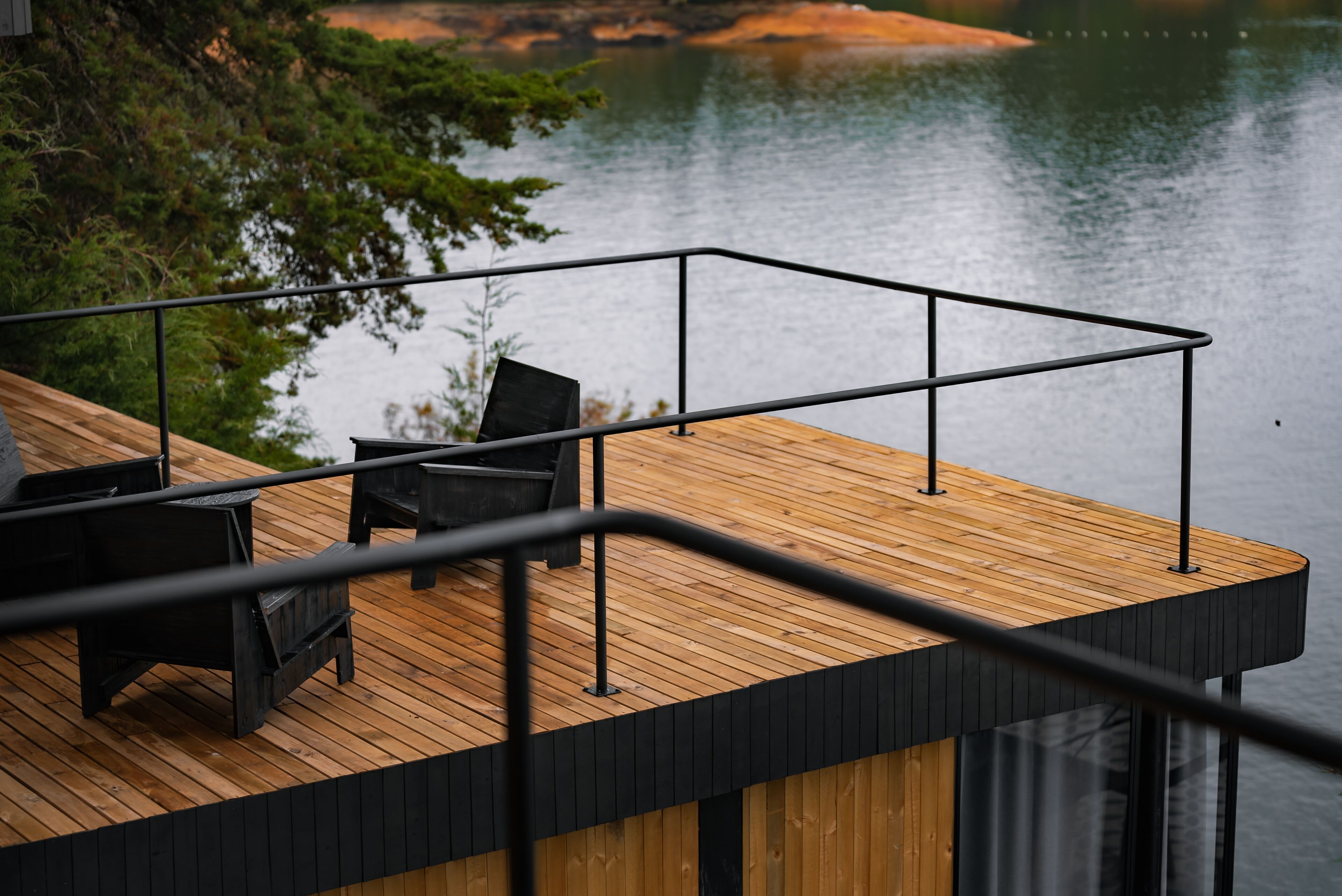 A wooden deck with a black railing, overlooking a calm lake and surrounded by greenery.
