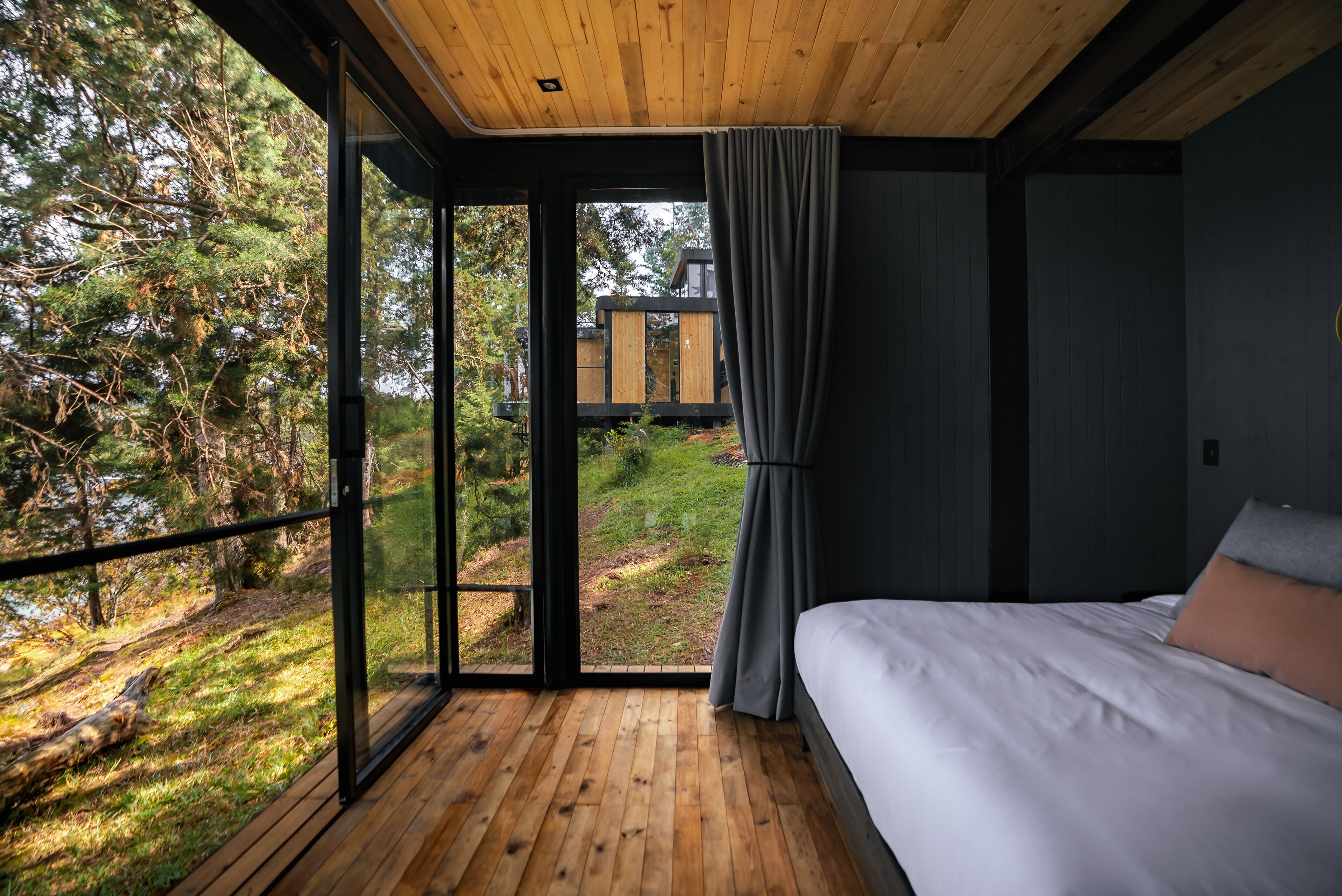 A bedroom with a wooden floor, a bed, a large window, and a view of a forest.