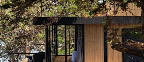 A modern cabin with a wooden deck, large glass windows, and a view of the surrounding forest.