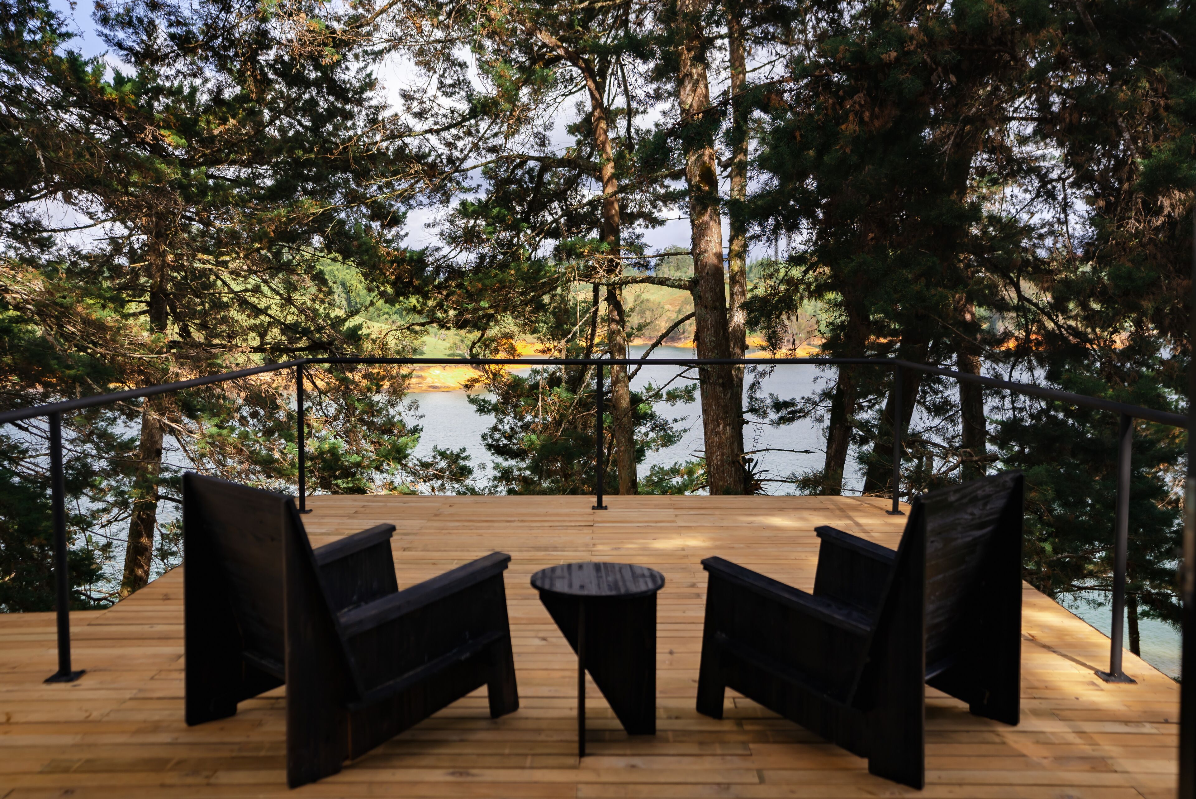 A wooden deck with two black chairs and a small round table, overlooking a lake surrounded by trees.