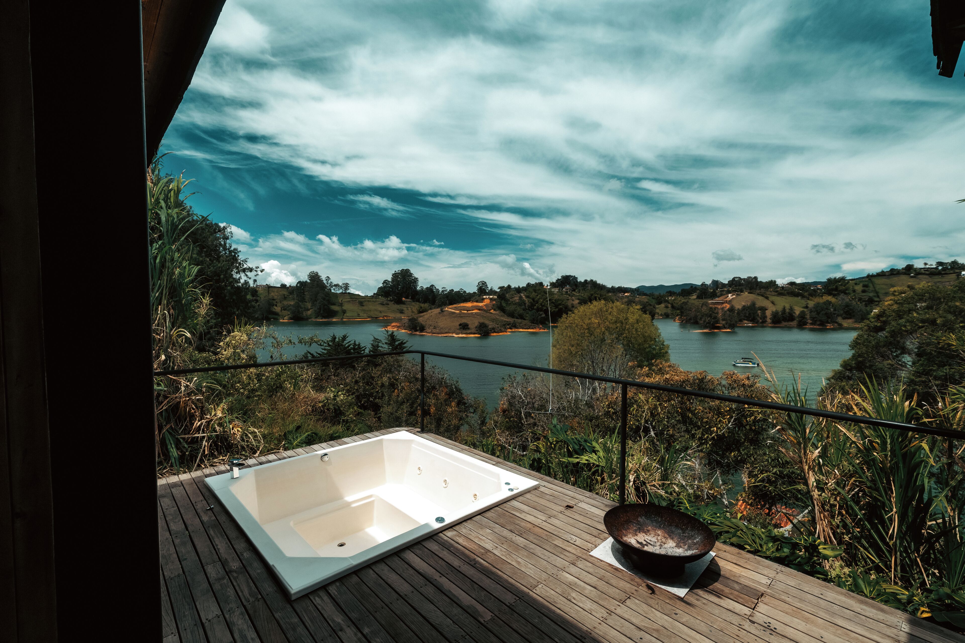 A wooden deck with a hot tub and a stone basin, overlooking a lake with boats and hills.