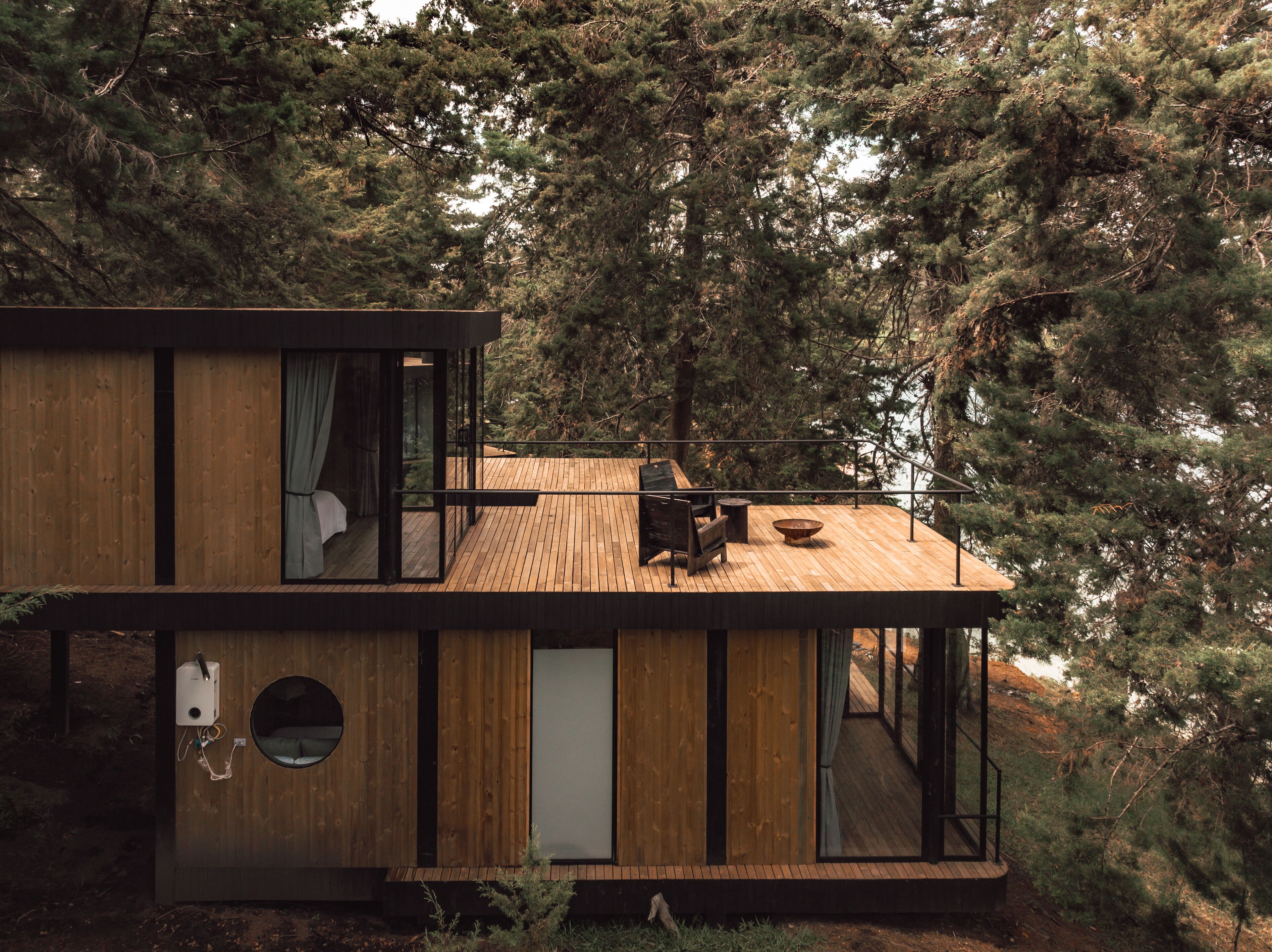 A modern cabin with a wooden exterior, large windows, and a deck overlooking a forested area.