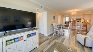 A spacious living room with a flat-screen TV, a dining area with a table and chairs, and a kitchen area with a sign reading "Emerald Isle". - Emerald Isle Vacation Rental w/ Resort Pool! (Emerald Isle)
