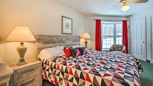 Una cama bien hecha con una colcha de estampado geométrico, dos lámparas de noche, un cabecero de madera, un cuadro en la pared, una ventana con cortinas rojas y un ventilador de techo.