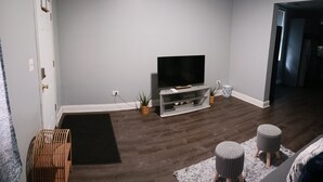 Una sala de estar con un televisor de pantalla plana sobre un mueble blanco, piso de madera oscura y alfombra blanca.