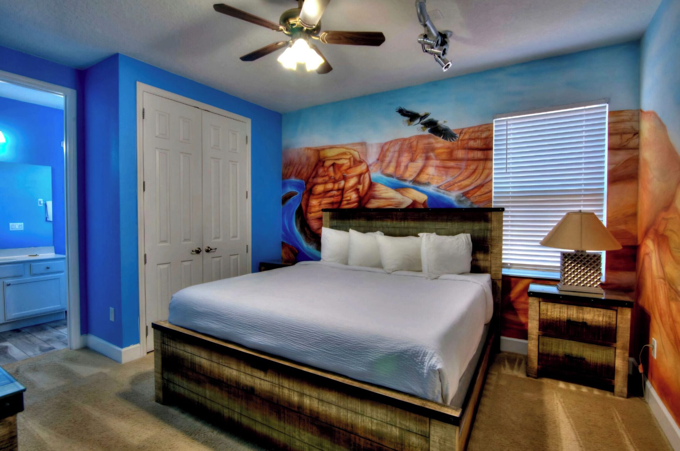 A bedroom with a bed, a ceiling fan, a nightstand with a lamp, and a mural on the wall.