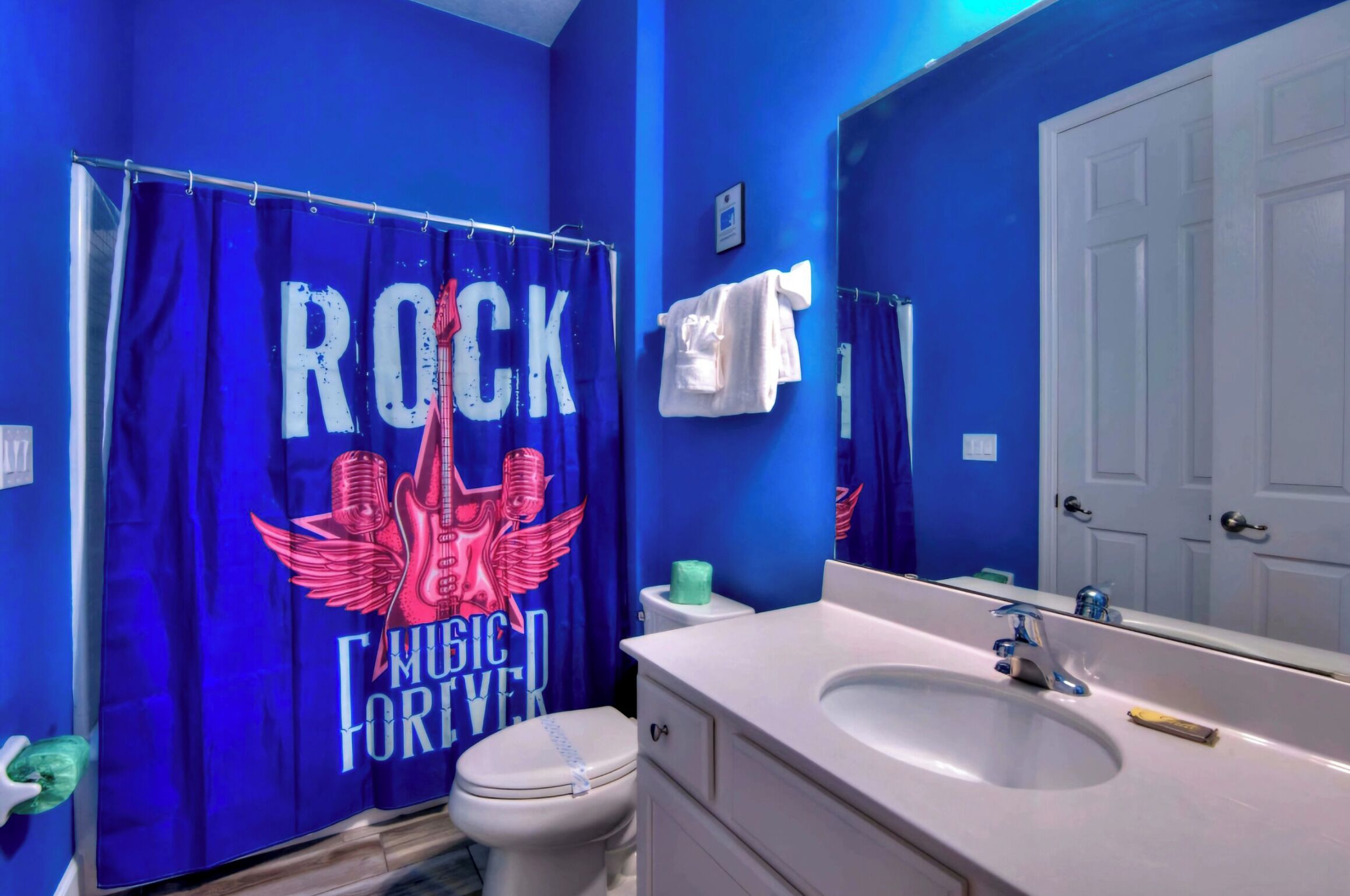 A bathroom with a blue wall, a white toilet, a sink with a mirror, and a shower curtain with "ROCK MUSIC FOREVER" text.