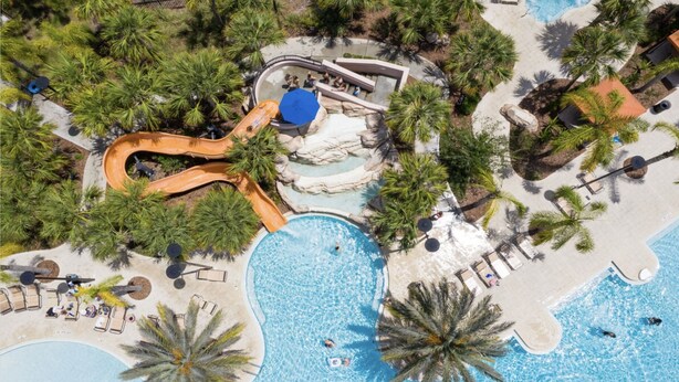Vista aérea de un resort con múltiples piscinas, un tobogán acuático y zonas de descanso rodeadas de palmeras.