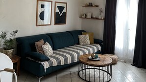 En stue med en grøn sofa, et rundt sofabord og en hvid stol. To billeder hænger på væggen, der er en plante, og en reol med pyngeting.