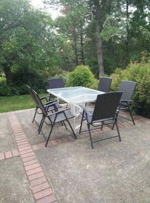 A patio with a glass-top table and four chairs, surrounded by greenery.