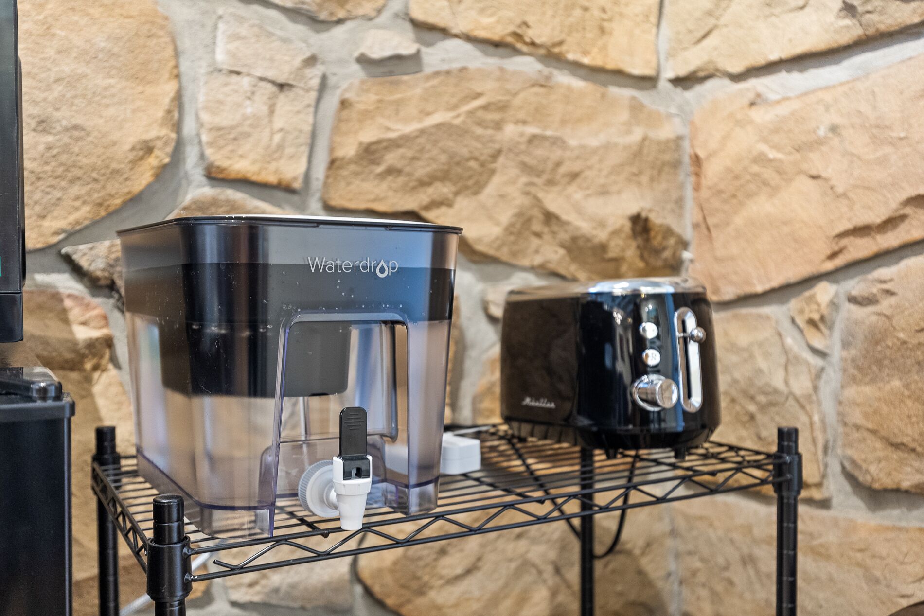A water dispenser and a toaster on a metal rack against a stone wall.