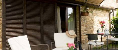A covered outdoor seating area with two chairs and a table, a stone wall, and a window with shutters.