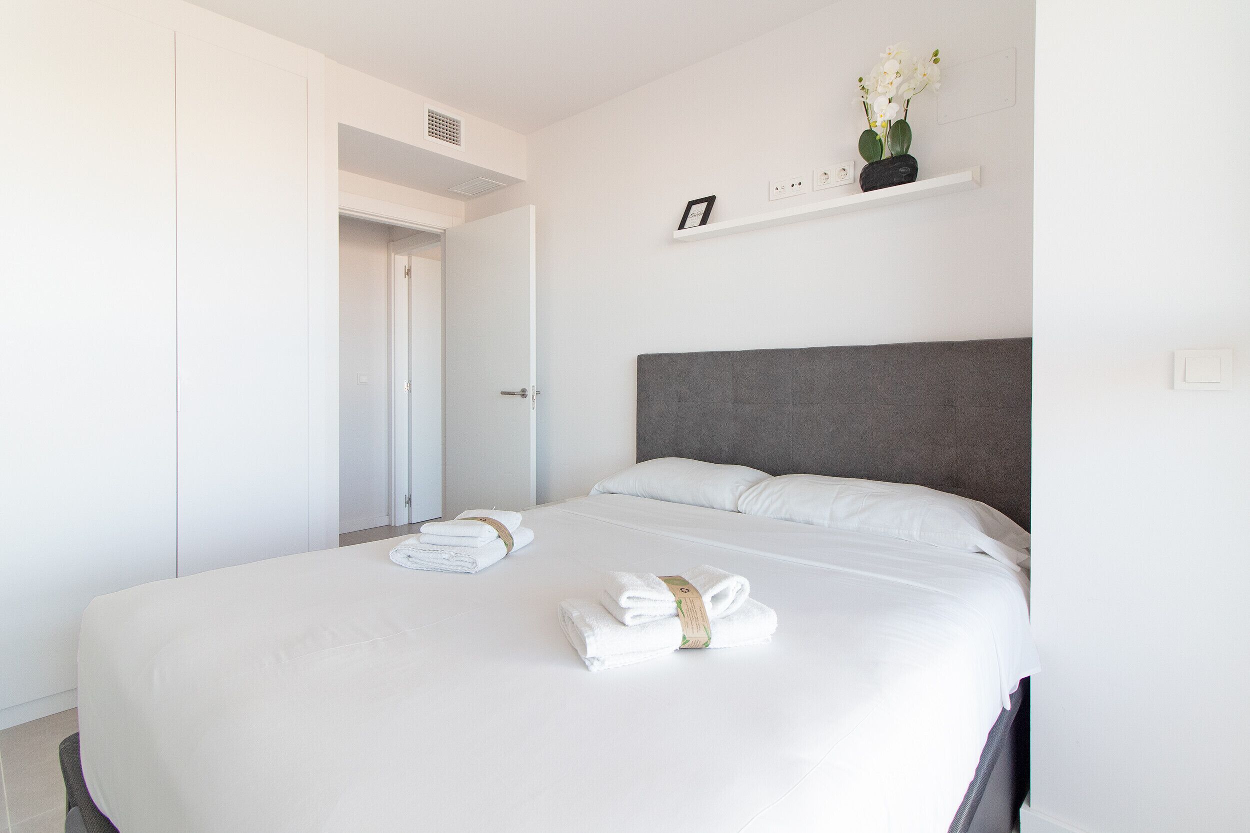 En snyggt bäddad säng med vita lakan och handdukar, en grå sänggavel och en liten hylla med en krukväxt.