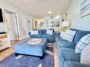 A living room with a blue sofa set, a coffee table, and a rug. There is a kitchen area in the background.