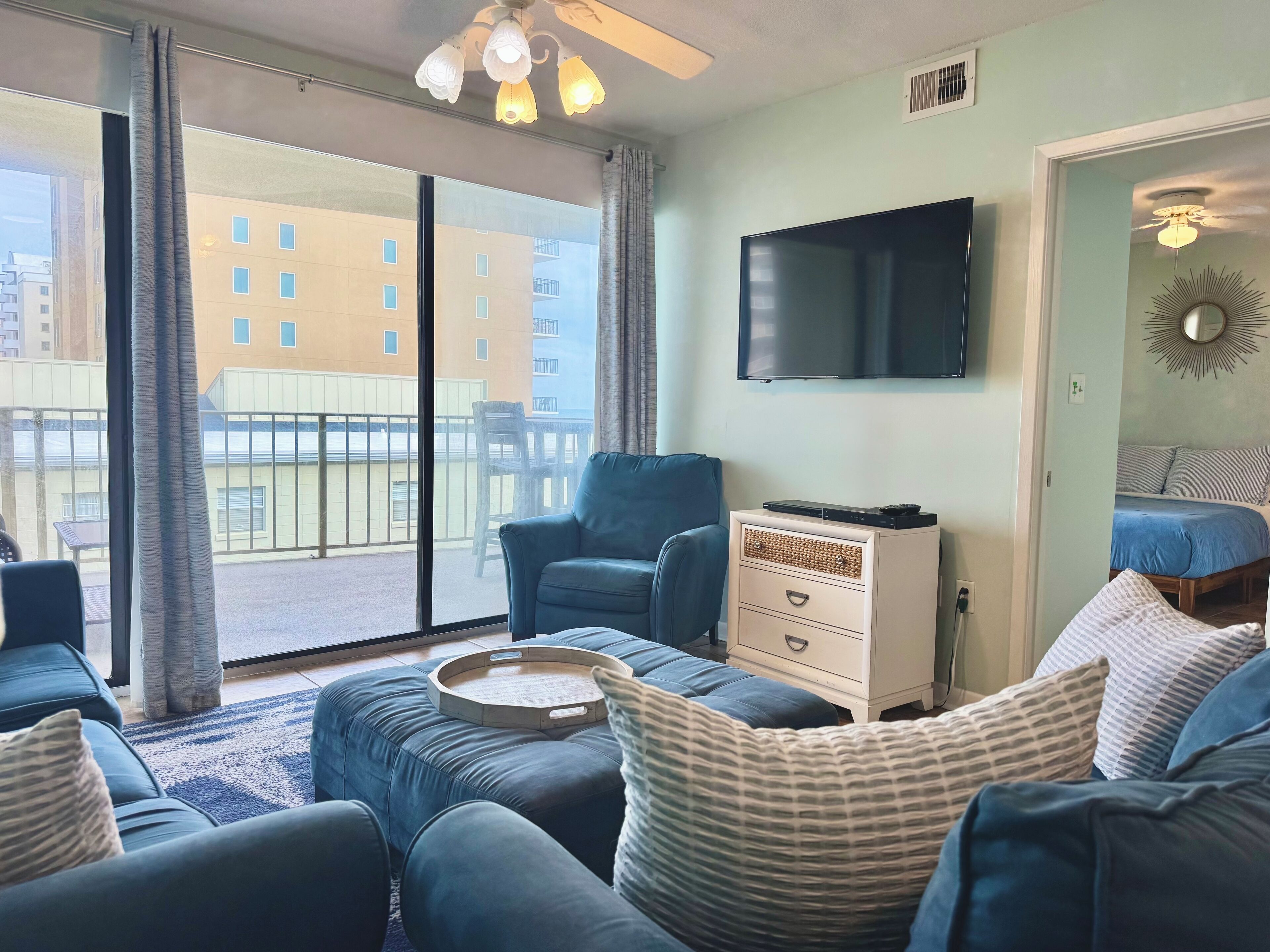 A living room with a large window, a flat-screen TV, a ceiling fan, and a bed with a blue bedspread.