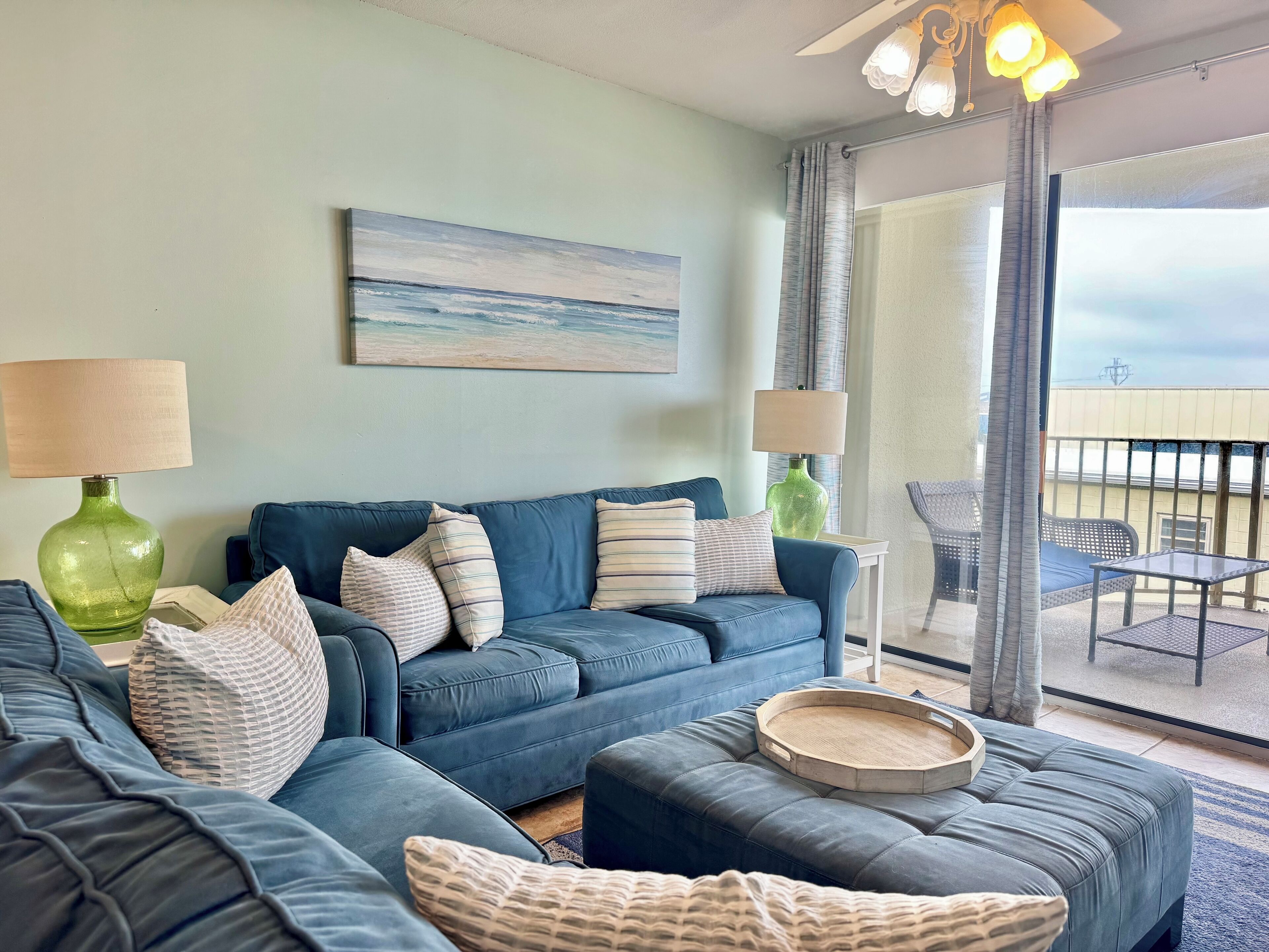 A modern living room with a blue sofa set, two side tables with lamps, a painting on the wall, and a balcony with outdoor furniture.