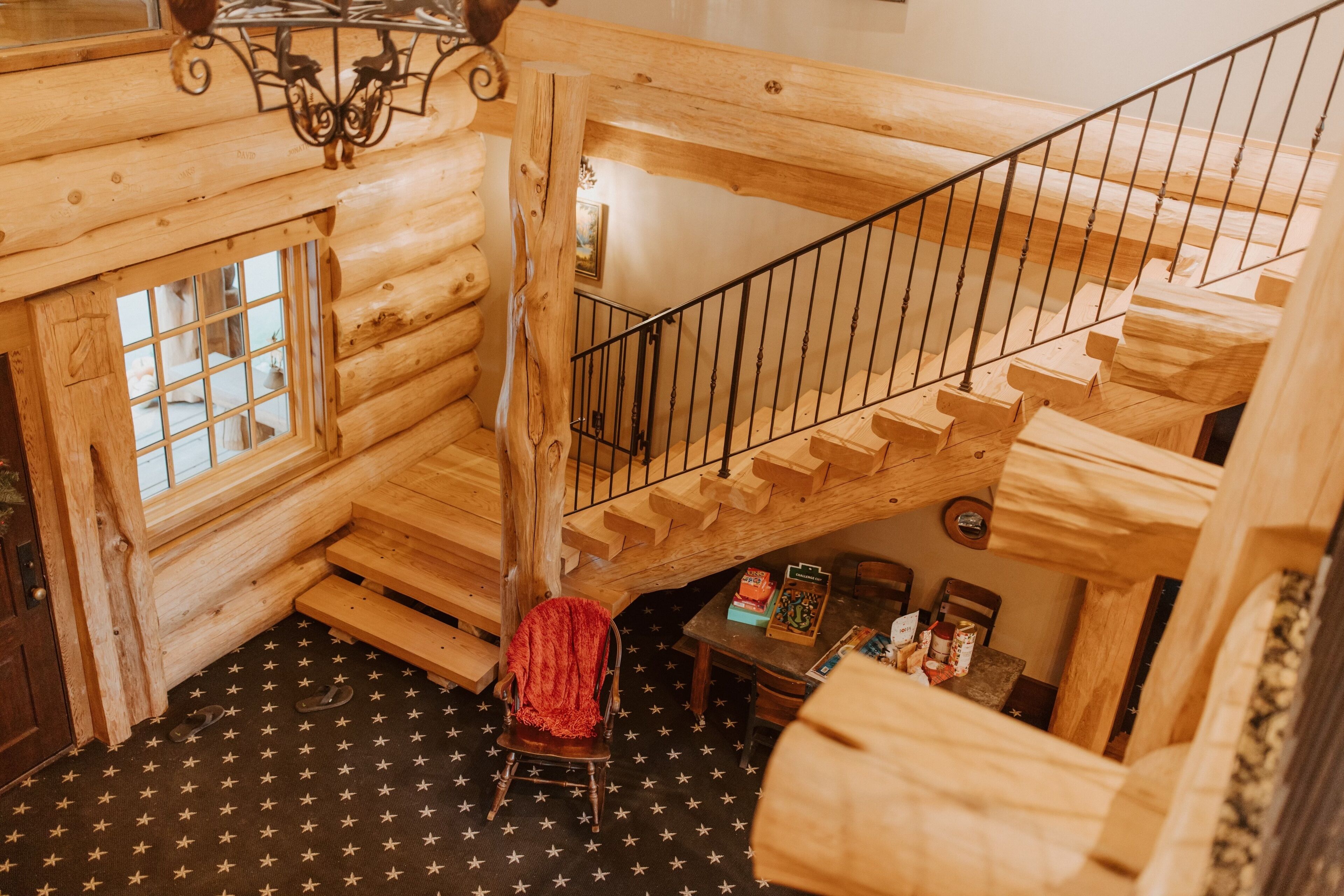 A wooden log cabin interior with a staircase, a chair, and a table with books and a lamp.