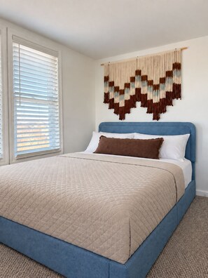 Un dormitorio con cama, cabecera, un adorno en la pared y una ventana con persianas.
