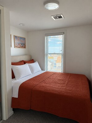 Une chambre à coucher avec un lit, une fenêtre avec des stores, un luminaire au plafond et un tableau accroché au mur.