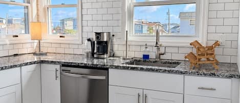 A modern kitchen with white cabinets, a stainless steel dishwasher, a coffee maker, and a view of the neighborhood through large windows.
