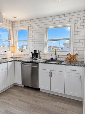 A modern kitchen with white cabinets, a stainless steel dishwasher, a coffee maker, and a view of the neighborhood through large windows. - Updated 5BR Home w/ Beach Badges 5 mins to the Beach (Stafford Township)