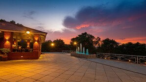 A covered outdoor area with a tiled floor, a building with a red roof, and a fountain. - Chinar Wood Resort Dapoli (Dapoli)