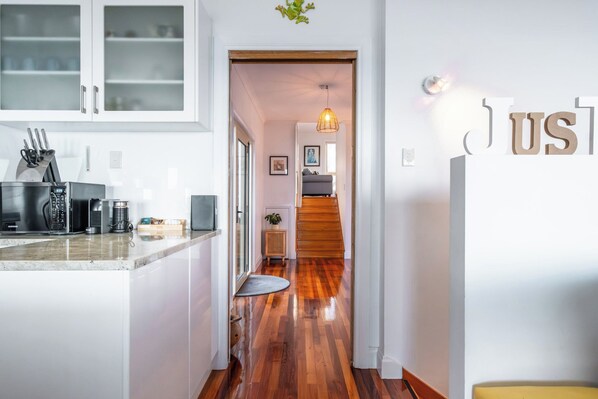 Una cucina moderna con mobili bianchi, un forno a microonde in acciaio inossidabile e una macchina per il caffè. Una scala in legno conduce a una zona giorno con un divano e una lampada.