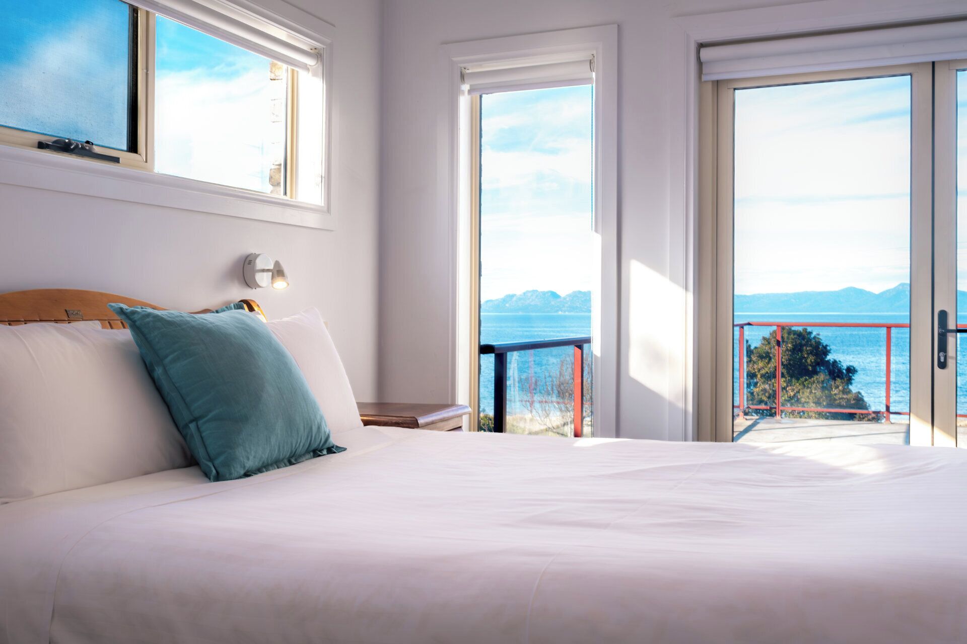 Un dormitorio con una cama grande, una mesita de noche, una lámpara de pared y vista al mar desde un balcón.