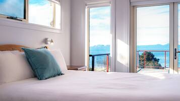 Un dormitorio con una cama grande, una mesita de noche, una lámpara de pared y vista al mar desde un balcón.