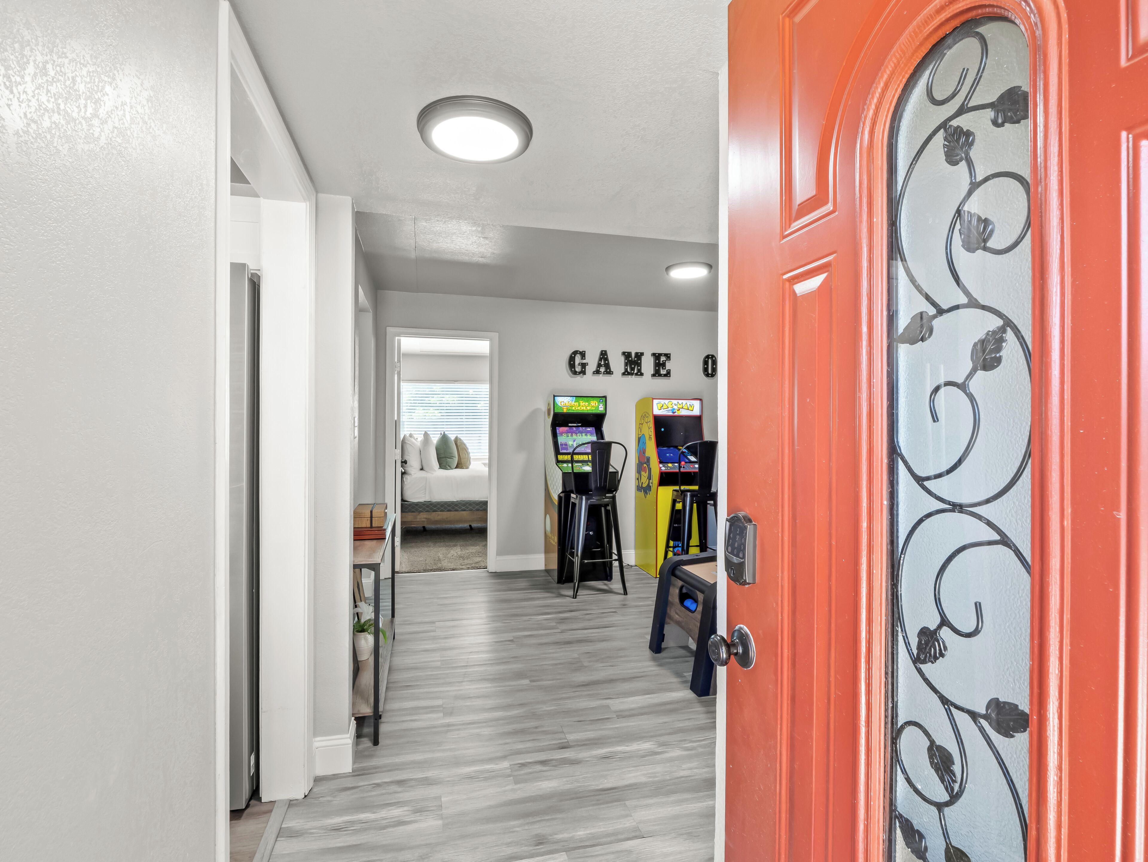 A hallway with a red door, a bed with pillows, and arcade games.