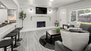 A modern living room with a white brick fireplace, a flat-screen TV mounted above it, a dining area with bar stools, and a large window with blinds.