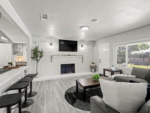 A modern living room with a white brick fireplace, a flat-screen TV mounted above it, a dining area with bar stools, and a large window with blinds.