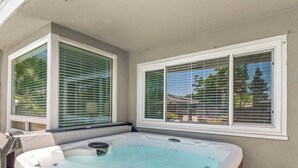 A hot tub with a wooden exterior, positioned adjacent to a house with white trim and green window blinds.