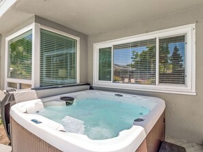 A hot tub with a wooden exterior, positioned adjacent to a house with white trim and green window blinds.