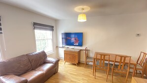 Un salon avec un canapé en cuir marron, une table à manger en bois avec des chaises, un téléviseur fixé au mur et un luminaire suspendu au plafond.