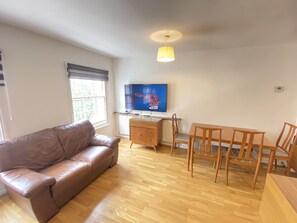 A living room with a brown leather sofa, wooden dining table with chairs, a TV mounted on the wall, and a ceiling light fixture.