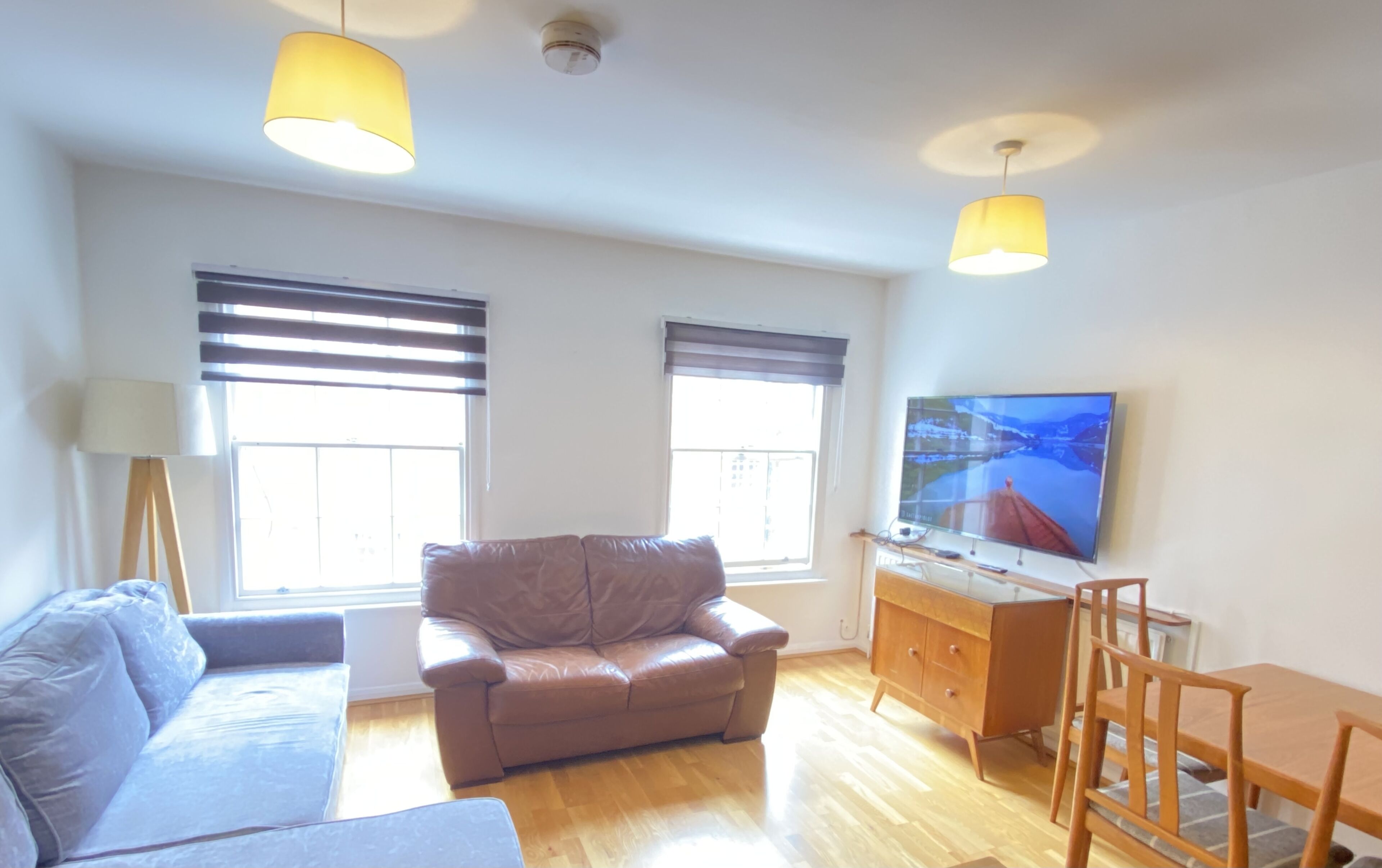 Ein Wohnzimmer mit braunem Ledersofa, einem blauen Sofa, einem Holzesstisch mit Stühlen, einem Fernseher an der Wand und zwei Fenstern mit Jalousien.