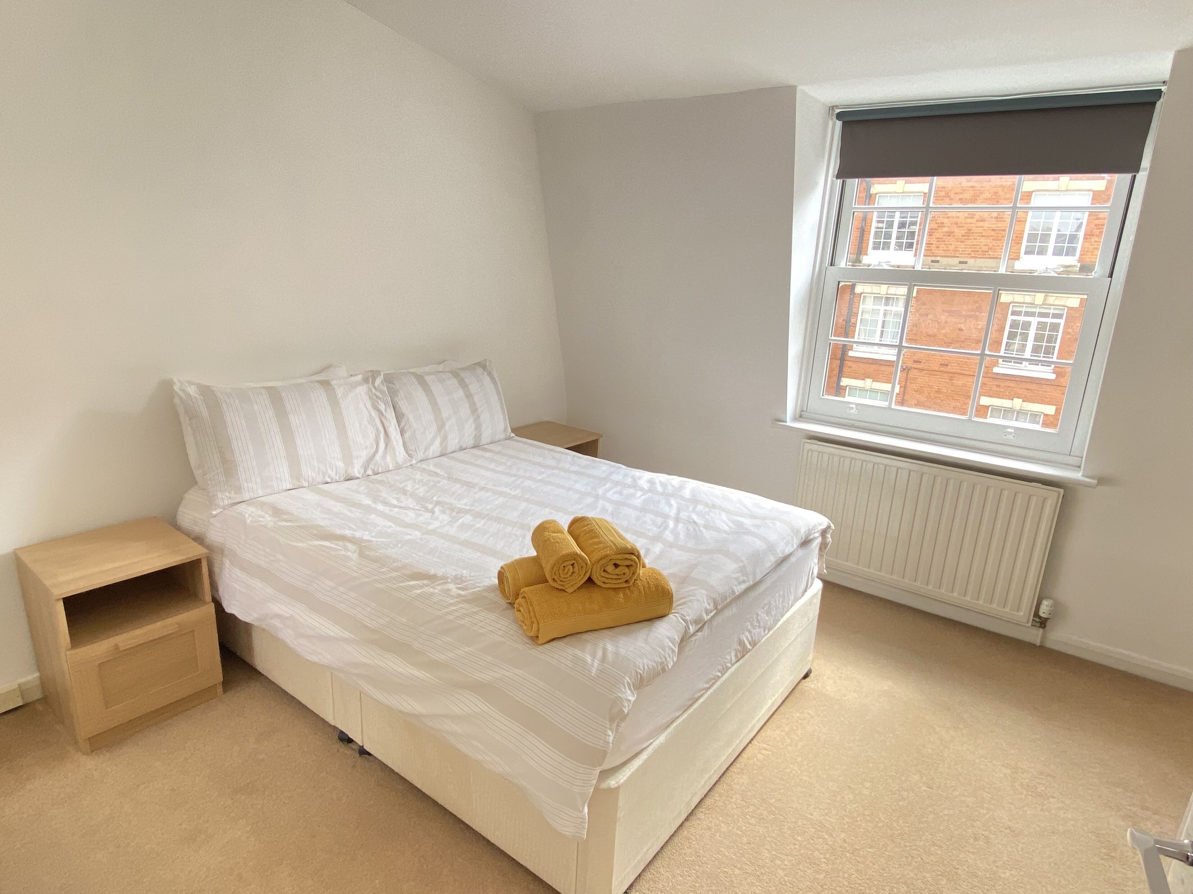 Slaapkamer met een enkel bed, een raam met uitzicht op gebouwen, een radiator en een klein houten nachtkastje.