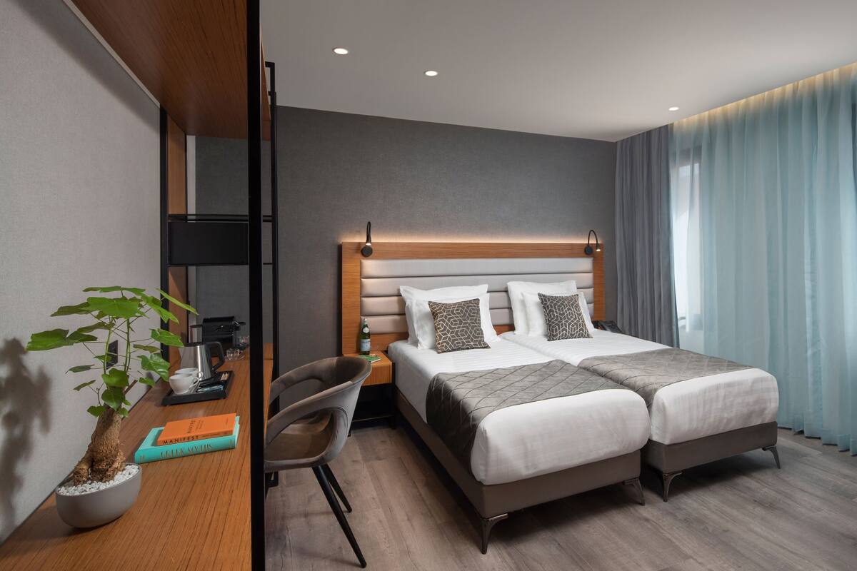 A hotel room with a bed, bedside table, chair, potted plant, and a view of the outside.
