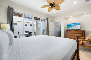 A bedroom with a large bed, a ceiling fan, a television, and a view of the outside.