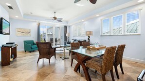 A spacious dining area with a glass table and wicker chairs, a ceiling fan, and large windows with blinds.