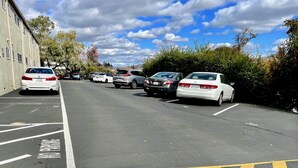 A parking lot with multiple cars parked in designated spaces, a building on the left, and trees in the background.