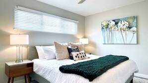 A bedroom with a bed, two bedside tables, a ceiling fan, a window with blinds, and a painting on the wall.
