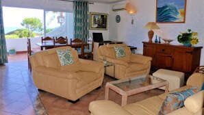A living room with two sofas, a coffee table, and a dining area with chairs and a table. There is a painting on the wall and a lamp on a cabinet. - Vilas Marrocha (Lagoa)