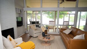 A spacious living room with a fireplace, a television, and a large window overlooking a deck. - Inks Lake Open Water and Pet friendly home (Burnet)