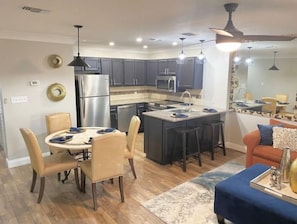 A modern kitchen with dark cabinets, stainless steel appliances, and a central island. There is a dining area with a round table and chairs. - Cozy condo in holiday island (Destin)